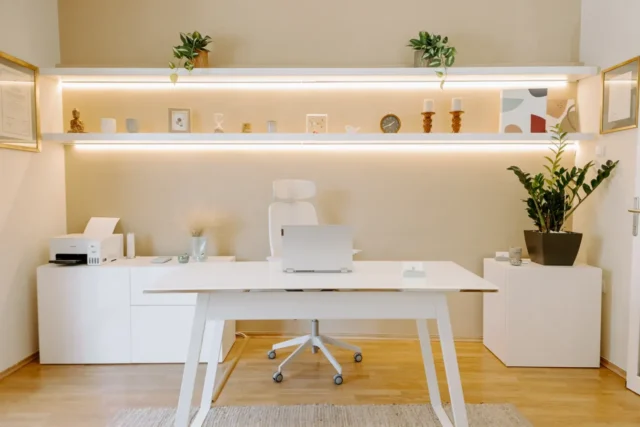 Μοντέρνο, μινιμαλιστικό γραφείο με λευκό γραφείο και καρέκλα. Ράφια με φυτά και διακοσμητικά στοιχεία, δημιουργώντας μια ήρεμη, οργανωμένη ατμόσφαιρα. Ξύλινο δάπεδο και απαλό φωτισμό.