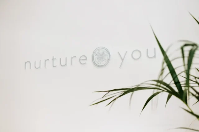 Το λογότυπο "nurture you" σε απαλό λευκό φόντο. Τα πράσινα φύλλα φυτών πλαισιώνουν εν μέρει την εικόνα, δημιουργώντας έναν ήρεμο, φροντιστικό τόνο.