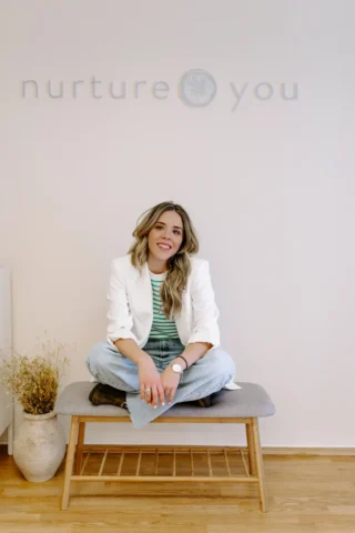 Χαμογελαστή γυναίκα με λευκό σακάκι και τζιν κάθεται οκλαδόν σε ένα παγκάκι σε ένα μινιμαλιστικό δωμάτιο με την επιγραφή «nurtureyou» στον τοίχο. Ένα βάζο με αποξηραμένα λουλούδια κοντά.