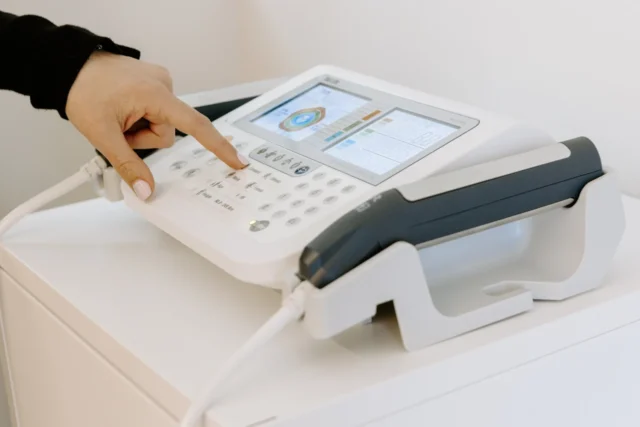 A hand with light pink nail polish presses a button on a white medical device for lipometry with a digital display and attached probes. The device sits on a white cabinet.
