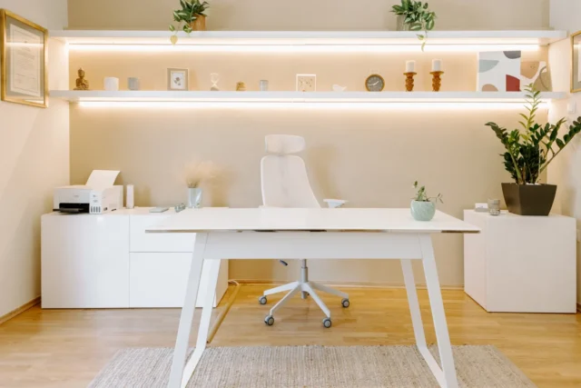 Μοντέρνο, μινιμαλιστικό γραφείο με λευκό γραφείο και καρέκλα. Ράφια με φυτά και διακοσμητικά στοιχεία, δημιουργώντας μια ήρεμη, οργανωμένη ατμόσφαιρα. Ξύλινο δάπεδο και απαλό φωτισμό.