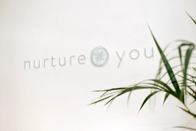 Το λογότυπο "nurture you" σε απαλό λευκό φόντο. Τα πράσινα φύλλα φυτών πλαισιώνουν εν μέρει την εικόνα, δημιουργώντας έναν ήρεμο, φροντιστικό τόνο.