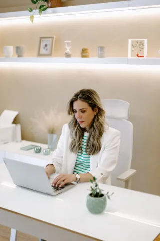 Μια γυναίκα με λευκό σακάκι εργάζεται σε έναν φορητό υπολογιστή σε ένα κομψό λευκό γραφείο, με ένα μικρό φυτό και διακοσμητικά στα ράφια. Το σκηνικό είναι φωτεινό και μοντέρνο.