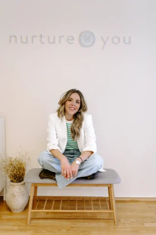 Χαμογελαστή γυναίκα με λευκό σακάκι και τζιν κάθεται οκλαδόν σε ένα παγκάκι σε ένα μινιμαλιστικό δωμάτιο με την επιγραφή «nurtureyou» στον τοίχο. Ένα βάζο με αποξηραμένα λουλούδια κοντά.