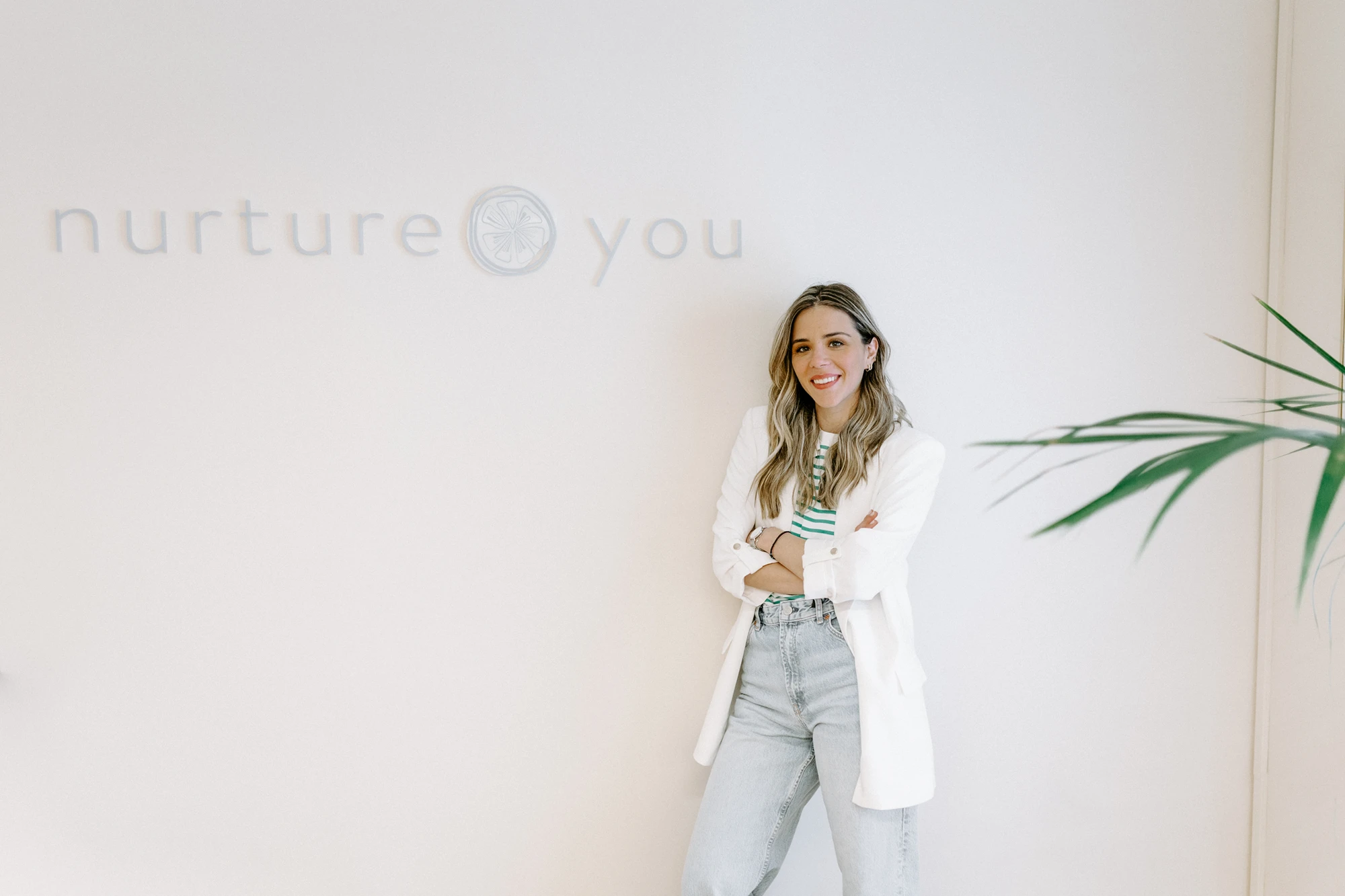 A woman in a white blazer and jeans stands in front of a wall with the words "nurture you," smiling with folded arms. A green plant is partially visible on the right.